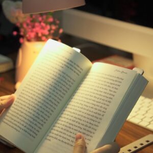 Hands holding an open book under a warm lamp on a desk, creating a cozy indoor reading environment.